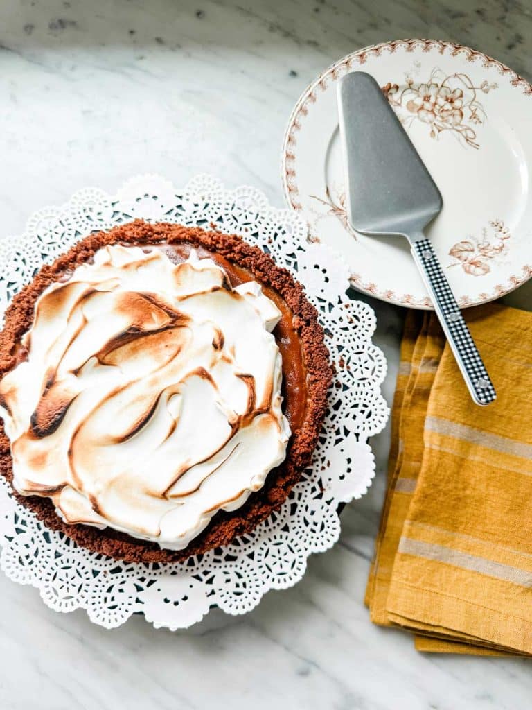 A chocolate meringue pie on a decorative doily, with a golden-brown topping, sits next to an ornate plate and a pie server on a yellow napkin on a marble surface, echoing the charm of a pumpkin pie recipe fit for any festive occasion.