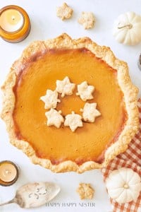 A pumpkin pie with a flaky crust is topped with leaf-shaped pastries, capturing the essence of a Costco Pumpkin Pie Recipe Copycat. It's beautifully arranged with miniature white pumpkins, lit votive candles, and a patterned cloth on a light surface.