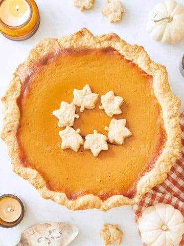 A pumpkin pie with a flaky crust is topped with leaf-shaped pastries, capturing the essence of a Costco Pumpkin Pie Recipe Copycat. It's beautifully arranged with miniature white pumpkins, lit votive candles, and a patterned cloth on a light surface.