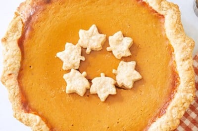 A pumpkin pie with a flaky crust is topped with leaf-shaped pastries, capturing the essence of a Costco Pumpkin Pie Recipe Copycat. It's beautifully arranged with miniature white pumpkins, lit votive candles, and a patterned cloth on a light surface.