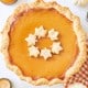 A pumpkin pie with a flaky crust is topped with leaf-shaped pastries, capturing the essence of a Costco Pumpkin Pie Recipe Copycat. It's beautifully arranged with miniature white pumpkins, lit votive candles, and a patterned cloth on a light surface.