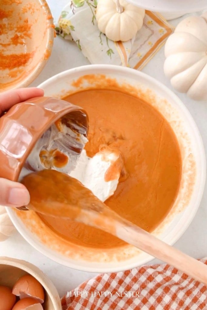 A hand pours whipped cream from a brown bowl into an orange pumpkin pie mixture in a white bowl. A spatula rests on the bowl's edge. The scene includes small white pumpkins and a checkered cloth on a light surface.