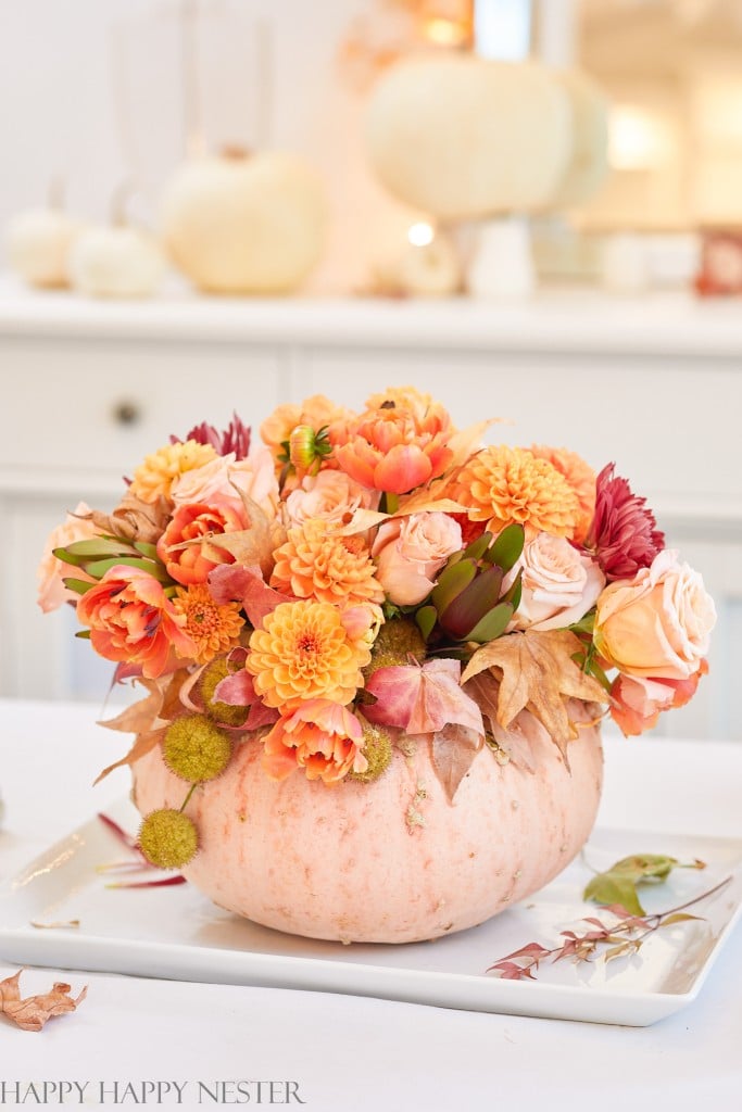 A pumpkin serves as an easy craft vase, filled with orange and peach flowers like roses and dahlias. Autumn leaves and buds complete the arrangement, elegantly displayed on a white tray. Subtle fall decorations grace the blurred background, enhancing the cozy autumnal vibe.