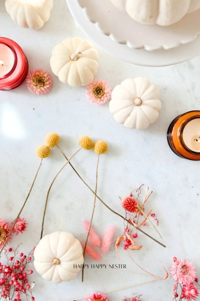 A flat lay of a fall-themed arrangement with small white pumpkins, pink flowers, yellow ball flowers, and candles on a light surface. The text "HAPPY HAPPY NESTER" is at the bottom.