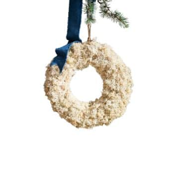 A decorative wreath made of dried, light-colored moss hangs on a dark blue ribbon. A small pine branch is visible above it, set against a plain white background.