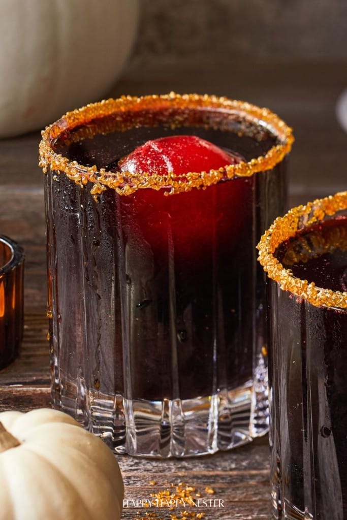 A dark black cocktail drink in a glass with a sugared rim features a large red spherical ice cube. A small white pumpkin is partially visible next to the glass on the wooden surface.