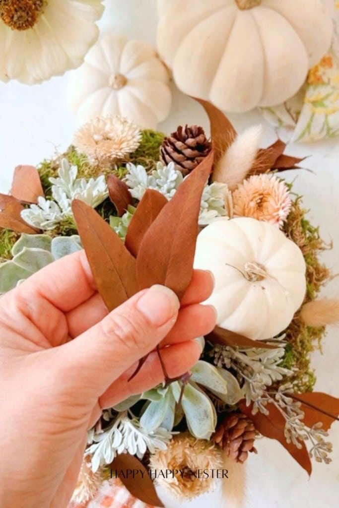 A hand holding two brown leaves over a fall-themed floral arrangement with succulent touches, white pumpkins, pinecones, and various rustic foliage. This DIY masterpiece captures the essence of autumn perfectly.