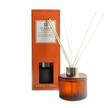 A ginger latte scented reed diffuser set, featuring an amber glass container with several reeds inserted. The matching rectangular box is displayed in the background, colored in warm orange tones with product details.