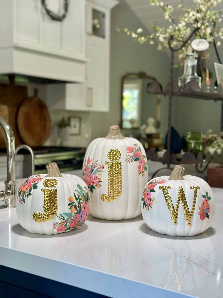 On a sleek white countertop in a modern kitchen, three DIY white pumpkins showcase floral designs and gold beaded letters "j," "j," and "w.