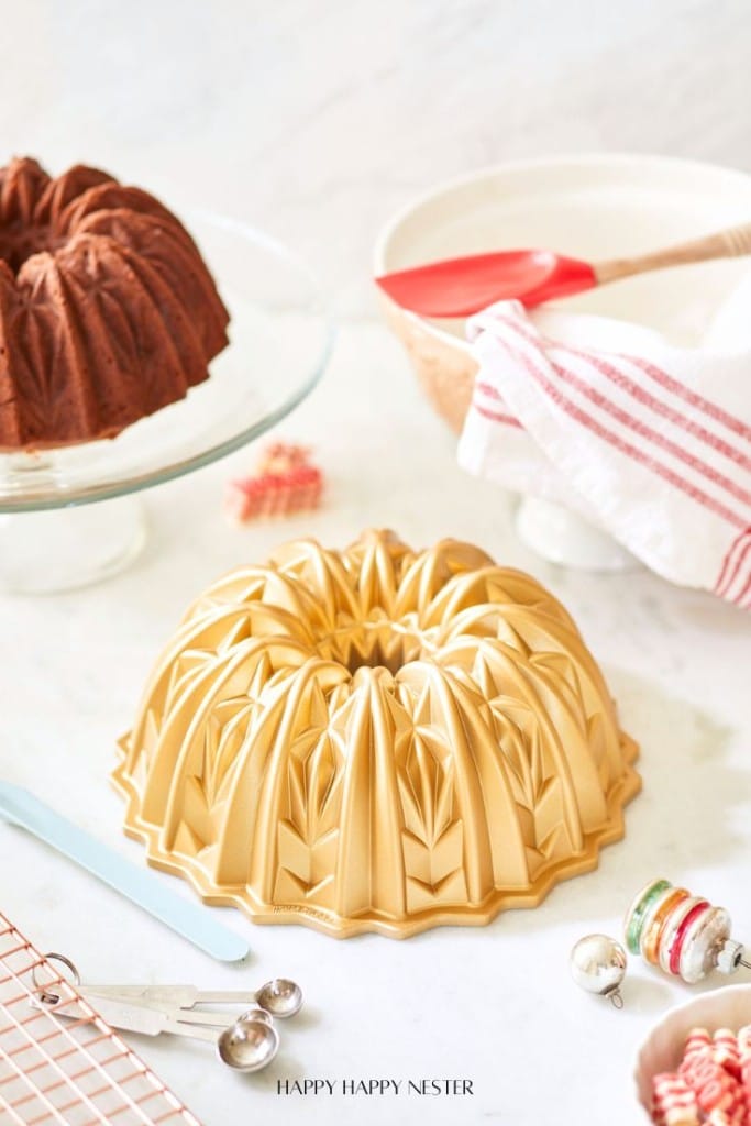 A decorative Bundt cake mold is displayed on a white surface alongside a tempting banana bread Bundt cake, made using oil, on a glass stand. Nearby are a red spatula, a striped cloth, measuring spoons, and a small whisk.