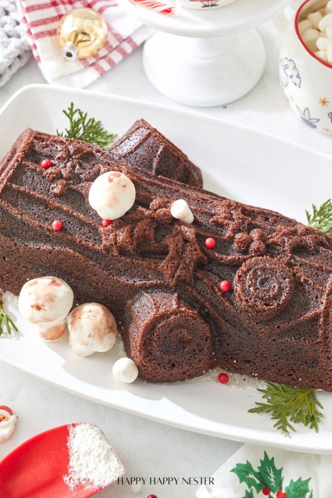 A Nordic Ware Yule Log cake, adorned with red berries and festive patterns, is surrounded by whimsical meringue mushrooms and lush greenery on a white platter.