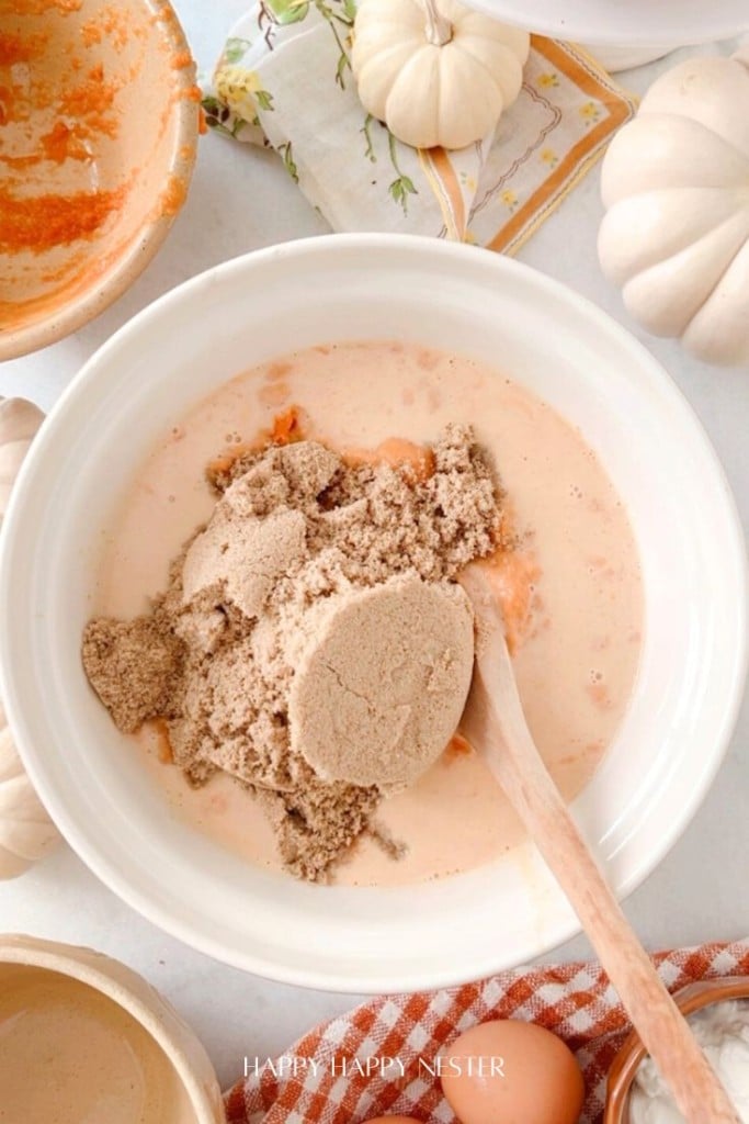 A white bowl brimming with a creamy pumpkin pie mixture, reminiscent of a Costco Pumpkin Pie Recipe Copycat, is topped with brown sugar. A wooden spoon rests in the mix. Surrounding the bowl are small white pumpkins, a napkin, and a partially filled glass of liquid.