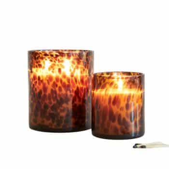 Two lit candles in amber glass holders with a speckled pattern. One is taller than the other. A partially burnt match lies nearby. The soft glow suggests a warm and cozy ambiance.