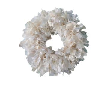 A decorative wreath made of soft, light-colored fabric ribbons, arranged in a fluffy and textured circular pattern against a plain white background.