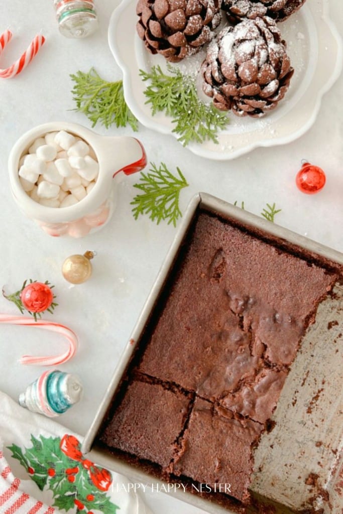 A tray of fudgy pinecone brownies, with a few pieces cut out, sits on a table. Nearby, a mug of marshmallows and chocolate pine cones dusted with powdered sugar are arranged on a plate. Festive ornaments and greenery adorn the scene, completing this Pinecone Brownies recipe setup.