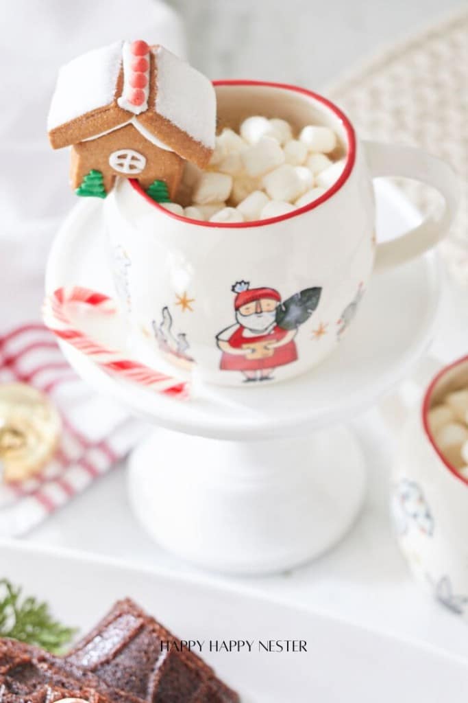 A festive mug filled with marshmallows sits on a white stand. The mug, featuring a Santa design, is adorned with a small gingerbread house and candy cane. Nearby, a red and white striped cloth adds to the holiday decor, reminiscent of cozy evenings baking Nordic Ware yule log recipes.