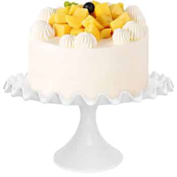 A white-frosted cake topped with cubes of yellow fruit, a single blueberry, and dollops of whipped cream rests on a ruffled white cake stand, capturing the essence of fall pies and cakes.