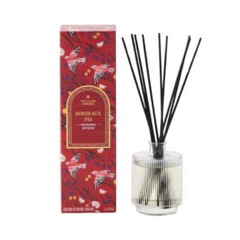 A fragrance diffuser with black reeds is displayed next to its decorative red box, labeled "Bordeaux Fig." The box has a floral pattern. The diffuser bottle is transparent, containing a light-colored liquid.