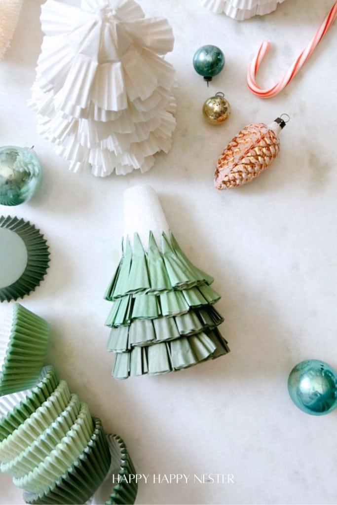 Festive scene with small paper tree decorations, green cupcake liners, a candy cane, and ornaments on a white surface.