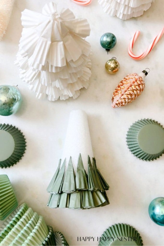 The table is a festive scene with craft materials, featuring a Paper Christmas Tree DIY project. A green paper cone takes center stage, resembling a Christmas tree, surrounded by white and green cupcake liners, small ornaments, and whimsical candy canes.