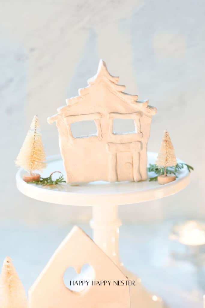 A whimsical air-dried clay house sits on a cake stand, surrounded by tiny bottle brush trees. The scene is light and playful, with soft lighting and a heart cutout at the base of the stand.