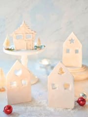 A wintry scene with minimalist gingerbread and air-dried clay houses alongside small Christmas trees on a snowy surface. One house sits on a white cake stand, surrounded by red and silver ornaments. Soft lighting creates a cozy holiday atmosphere.