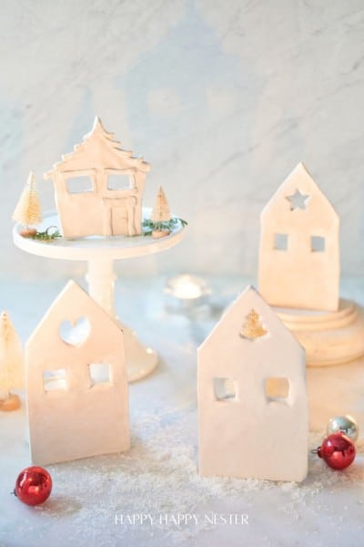 A wintry scene with minimalist gingerbread and air-dried clay houses alongside small Christmas trees on a snowy surface. One house sits on a white cake stand, surrounded by red and silver ornaments. Soft lighting creates a cozy holiday atmosphere.