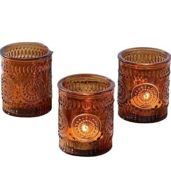 Three brown glass candle holders with intricate circular designs, each containing a lit candle. The warm glow illuminates the ornate patterns on the glass, creating a cozy ambiance.