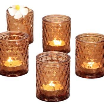 Five brown glass candle holders with a honeycomb pattern are lit with tealight candles inside. The back left holder is topped with a white and yellow plumeria flower.