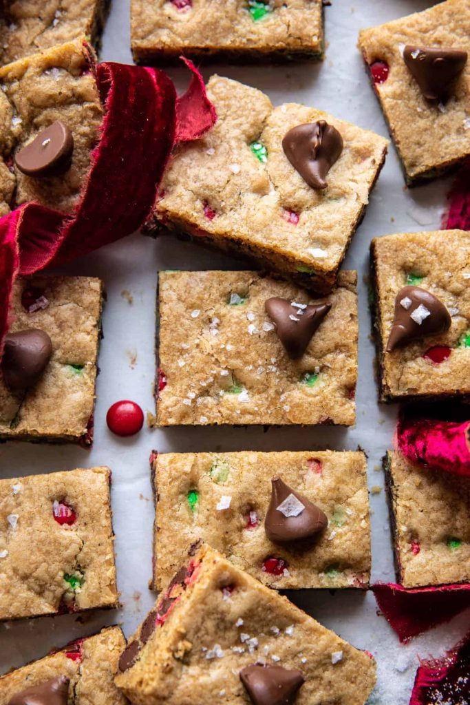 Close-up of cookie bars with chocolate pieces and colorful candy bits inside. Some are sprinkled with sea salt, creating the perfect Christmas treats. A red velvet ribbon weaves among the cookies, adding a festive touch.