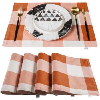 A set of four folded orange and white checkered placemats with one unfolded on top. On the open placemat is a white plate featuring a black geometric design, accompanied by a black and gold fork and spoon.