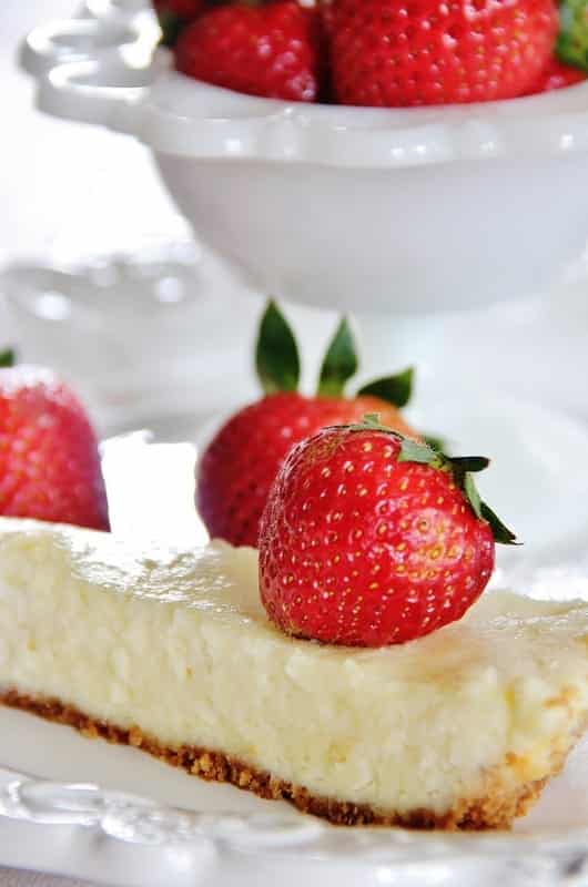 A slice of cheesecake, offering cake inspiration, is topped with a fresh strawberry on a white plate. In the background, a bowl brimming with ripe strawberries adds to the tempting display.