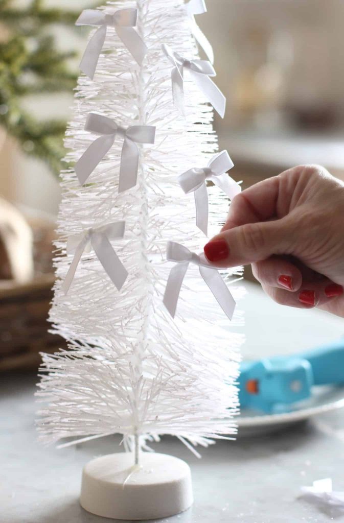 A hand with red nail polish decorates a small white bottlebrush Christmas tree with elegant white bows, an ideal piece for holiday craft ideas. The tree sits on a gray surface, while a blue tool is artfully blurred in the background.