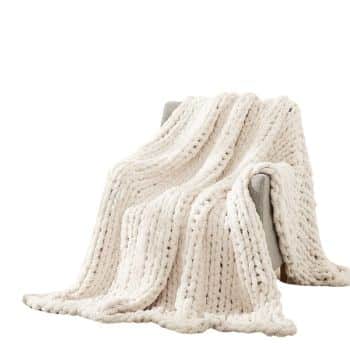 A large, soft cream-colored chunky knit blanket draped over a gray chair against a plain white background.