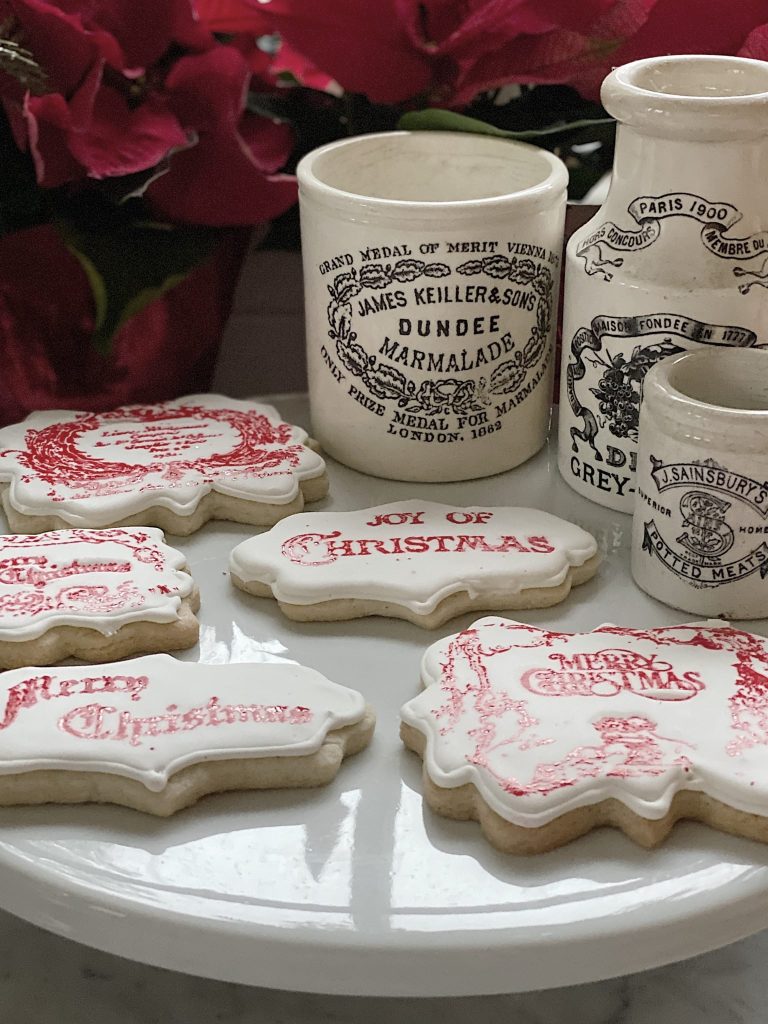 A collection of the best Christmas cookies with festive messages is displayed on a white plate. Behind them, vintage-style ceramic jars boast decorative labels. Red poinsettias are partially visible in the background.