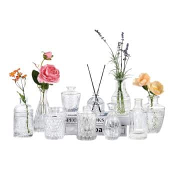 A collection of clear glass vases filled with colorful flowers, including pink roses, orange blossoms, and lavender, arranged on books. Each vase has a unique design, adding to the decorative ensemble.