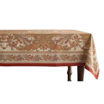 A dining table with a patterned tablecloth featuring intricate orange and beige paisley designs on a light background, with a decorative border. The wooden table leg is visible at the lower right corner.