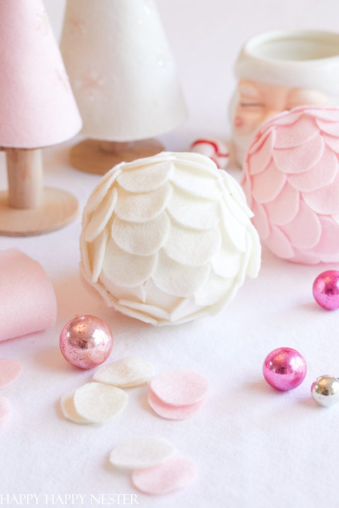 A cozy holiday scene featuring pink and white felt ornaments, some shaped like balls with petal patterns. Decorative trees and a Santa mug with rosy cheeks sit in the background, surrounded by small pink and silver baubles—perfect inspiration for Christmas crafts for adults.