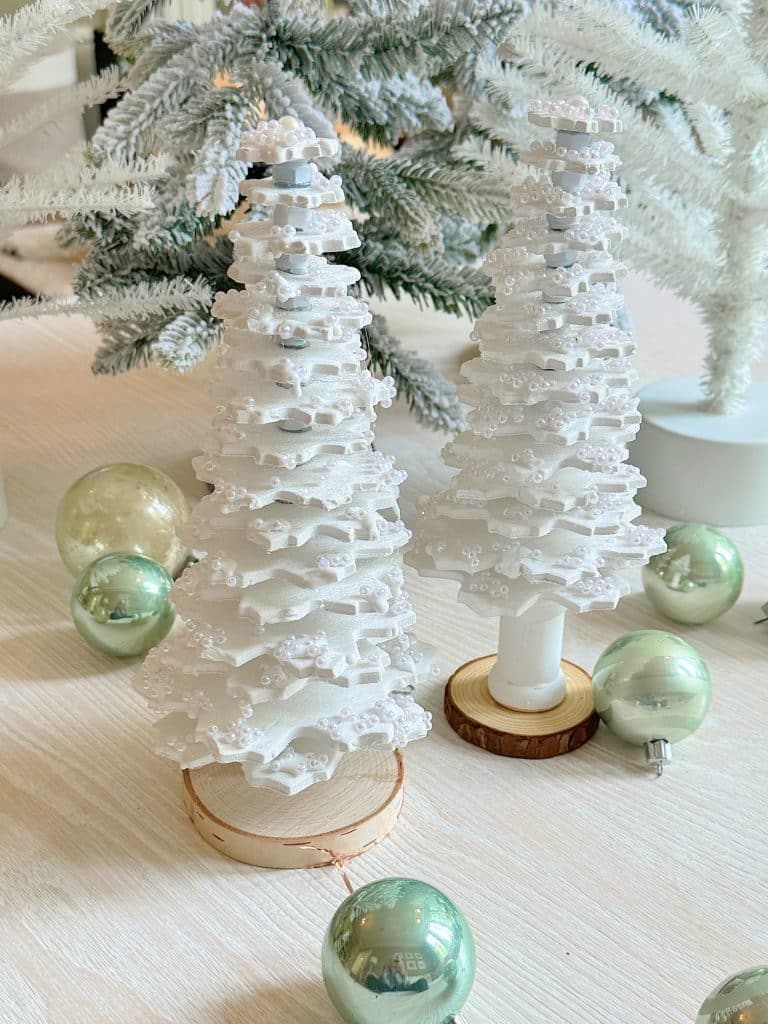 White decorative Christmas trees, perfect for Christmas crafts for adults, are adorned with textured layers and stand on circular wooden bases. They are surrounded by pale green ornaments on a light wood surface, alongside a frosted Christmas tree in the background.