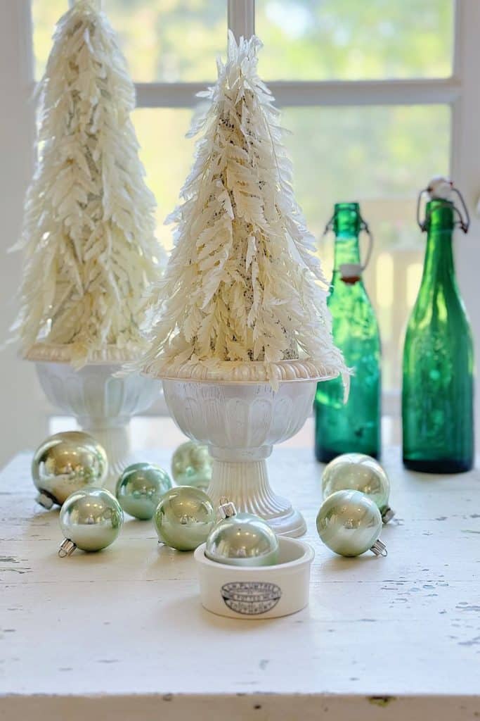 A festive scene with two white decorative Christmas trees in vases on a table, surrounded by green and silver ornaments. A subtle touch of whimsy is added with a paper Christmas tree centerpiece. Two green glass bottles stand in the background, illuminated by light streaming through a window.