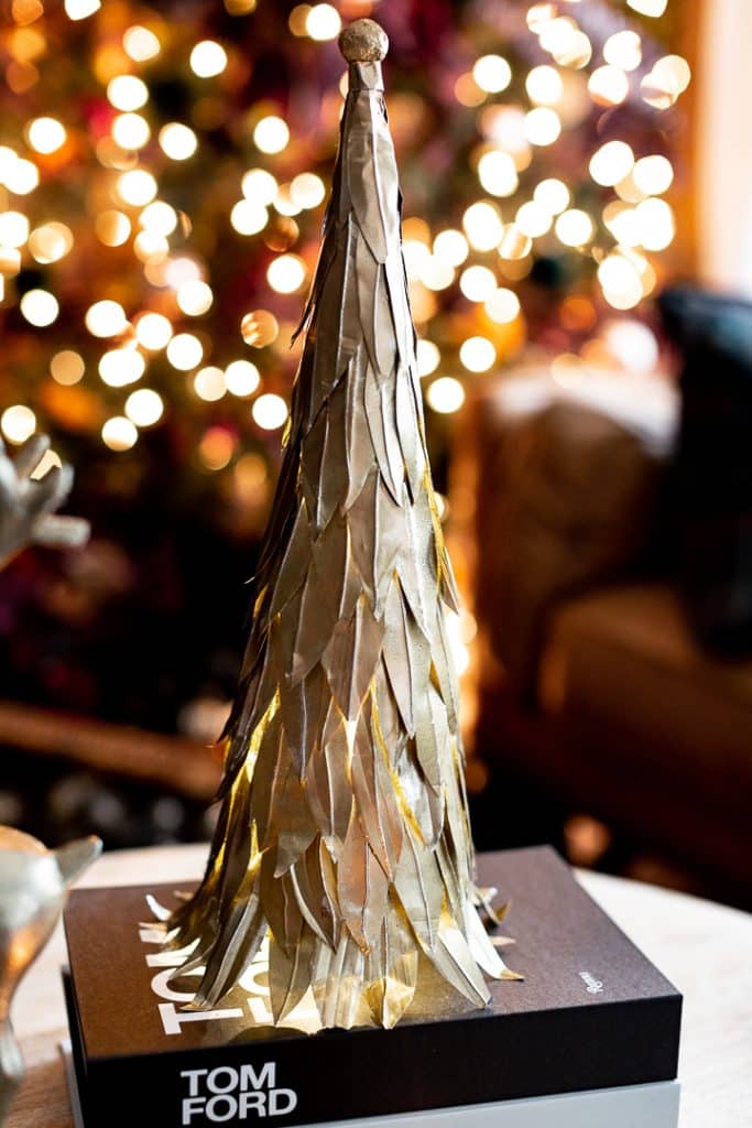 A decorative, gold-colored tree sculpture stands on a Tom Ford book, echoing the form of a paper Christmas tree. In the softly blurred background, a lit Christmas tree adds warmth. The sculpture's slender shape resembles a stylized tree formed from layered metal leaves.