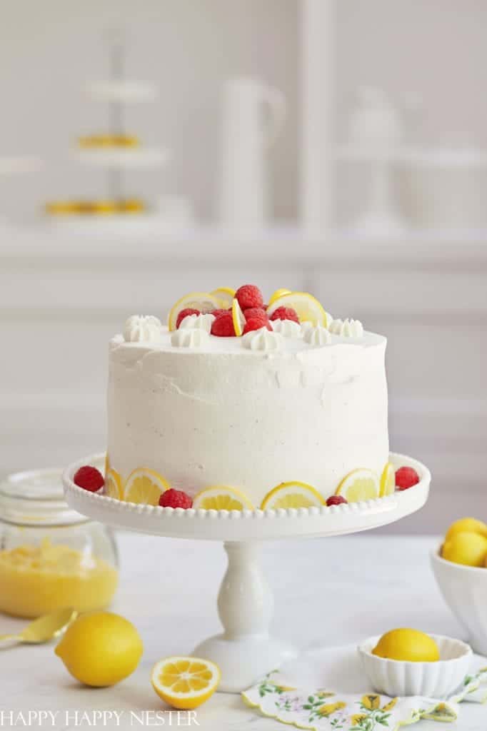 A beautifully decorated white cake on a pedestal stand, topped with raspberries and cream swirls, serves as perfect cake inspiration. Lemon slices and raspberries are artfully arranged around the base. In the blurred background, lemons, a jar of curd, and a tiered stand complete the elegant setting.