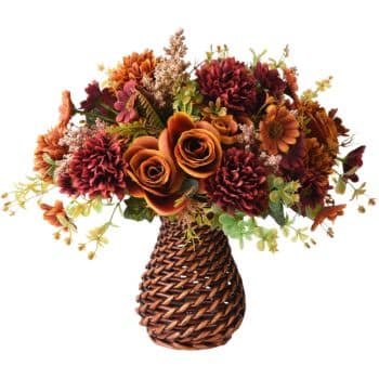 A woven vase holds a vibrant autumn bouquet featuring orange roses, red and burgundy chrysanthemums, and green foliage. The rich colors evoke a warm fall atmosphere.