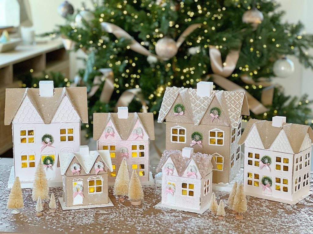 A collection of decorative gingerbread houses with white icing and small wreaths is displayed on a wooden table dusted with powdered sugar. For those seeking holiday craft ideas, miniature trees surround the houses, and a Christmas tree with lights and ornaments twinkles in the background.