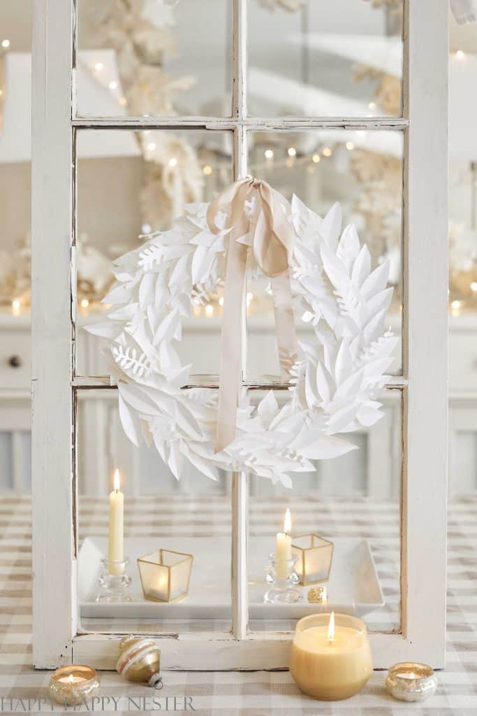 A white paper wreath with ribbon hangs on a vintage windowpane, embodying the charm of holiday craft ideas. Below, candles flicker in glass holders, surrounded by small decorations, creating a cozy, festive atmosphere with soft lighting and reflections.