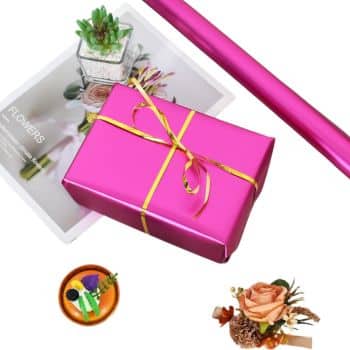 A pink gift box with a gold ribbon on a floral book, alongside a pink wrapping paper roll, a succulent plant, and flower decorations.