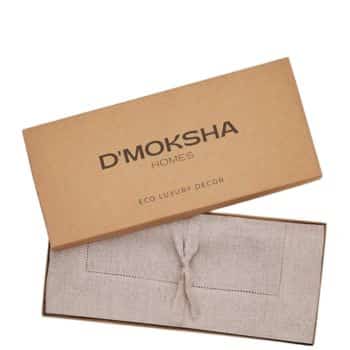 A cardboard box with "D'Moksha Homes" and "Eco Luxury Décor" printed on the lid. The box is open, revealing a folded beige fabric tied with a string inside.