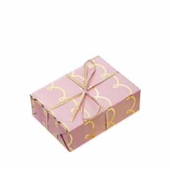 A small rectangular gift box wrapped in pink paper with gold decorative patterns. It is tied with a thin, shiny gold ribbon in a neat bow.