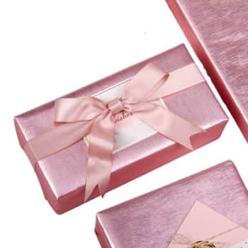 A rectangular gift box wrapped in pink metallic paper with a matching pink satin ribbon tied in a bow. The box is placed on a white surface among other wrapped gifts.