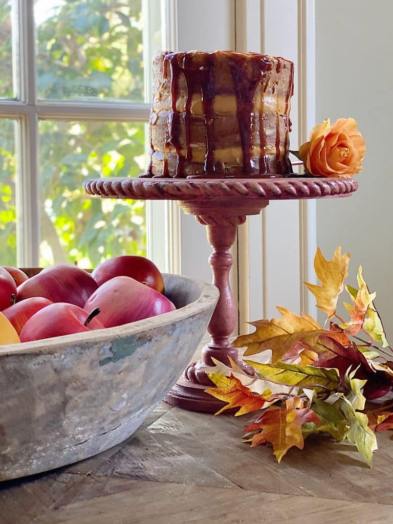 A caramel-drizzled cake on a red pedestal serves as delightful cake inspiration, with an orange rose beneath a sunlit window. A bowl of red apples and autumn leaves rests nearby on a wooden table, bathed in natural light.